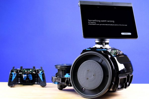 A partially disassembled Amazon Astro sits on a wooden desktop in front of a blue backdrop. The screen displays the text "Something went wrong"